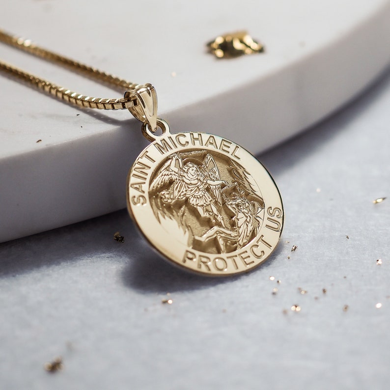 May include: Gold pendant necklace with a round charm featuring an image of Saint Michael the Archangel. The charm has the words "Saint Michael Protect Us" inscribed around the edge.