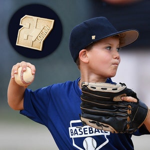 K&ouml;nnte beinhalten: Ein junger Baseballspieler tr&auml;gt ein blaues Trikot mit dem Wort "BASEBALL" darauf, eine marineblaue Baseballkappe mit wei&szlig;em Logo und einen schwarzen Baseballhandschuh. Der Spieler wirft einen wei&szlig;en Baseball mit roten N&auml;hten. Eine goldene Nummer 27 mit dem Namen "LOGAN" ist im Hintergrund.