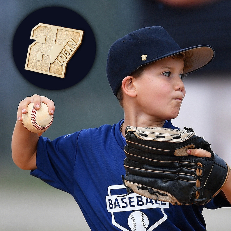 K&ouml;nnte beinhalten: Ein junger Baseballspieler tr&auml;gt ein blaues Trikot mit dem Wort "BASEBALL" darauf, eine marineblaue Baseballkappe mit wei&szlig;em Logo und einen schwarzen Baseballhandschuh. Der Spieler wirft einen wei&szlig;en Baseball mit roten N&auml;hten. Eine goldene Nummer 27 mit dem Namen "LOGAN" ist im Hintergrund.