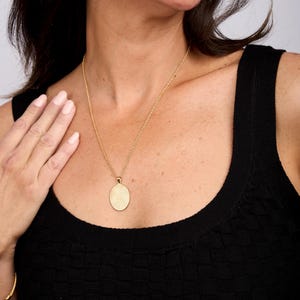 Puede incluir: Un collar de cadena de oro con un gran colgante ovalado. El colgante es liso y tiene una superficie ligeramente texturizada.