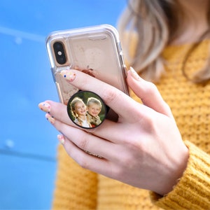K&ouml;nnte beinhalten: Ein schwarzer Handy-Griff mit einem runden Fotoeinsatz, der zwei l&auml;chelnde Kinder zeigt. Der Fotoeinsatz ist an einer durchsichtigen Handyh&uuml;lle befestigt.