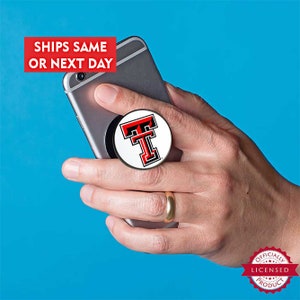 May include: A white phone pop socket with a red and black Texas Tech University logo. The logo features a large red "T" with a black outline and a smaller black "T" inside the red "T". The pop socket is attached to a silver smartphone.
