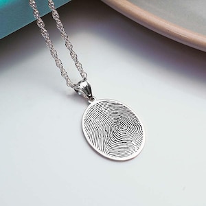 Puede incluir: Un collar de plata con un colgante que presenta un dise&ntilde;o detallado de huella digital. El colgante tiene forma ovalada y cuelga de una delicada cadena de plata.