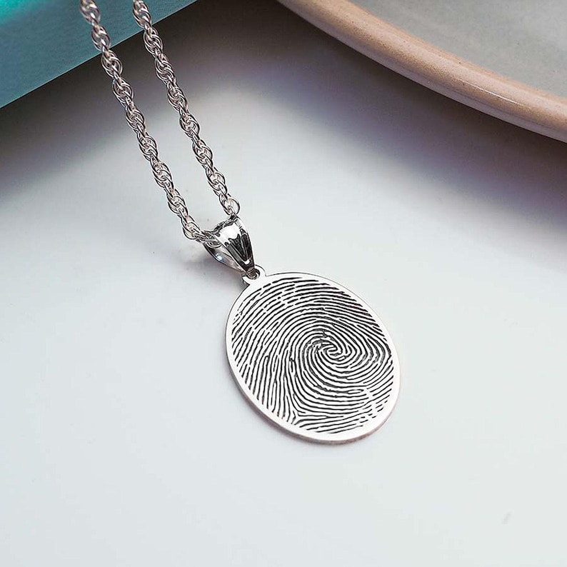 Puede incluir: Un collar de plata con un colgante que presenta un dise&ntilde;o detallado de huella digital. El colgante tiene forma ovalada y cuelga de una delicada cadena de plata.