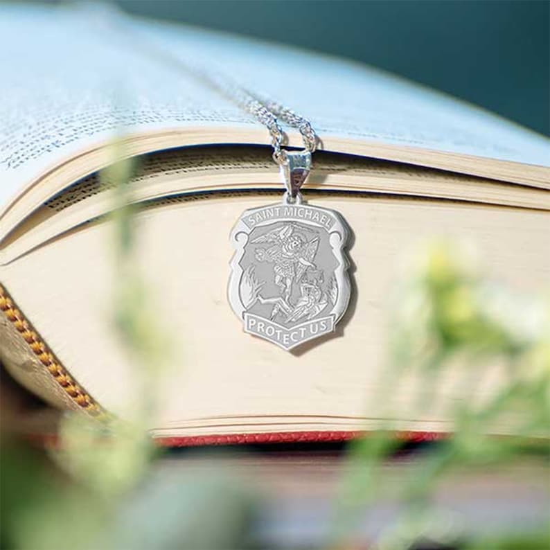 K&ouml;nnte beinhalten: Silberne Halskette mit einem Anh&auml;nger in Form eines Schildes, der ein Bild des Erzengels Michael zeigt. Der Anh&auml;nger tr&auml;gt die Inschrift "Saint Michael Protect Us".