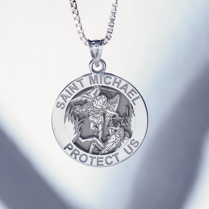 May include: Silver pendant necklace featuring a round charm with a depiction of Saint Michael slaying a demon. The charm is inscribed with "Saint Michael Protect Us".