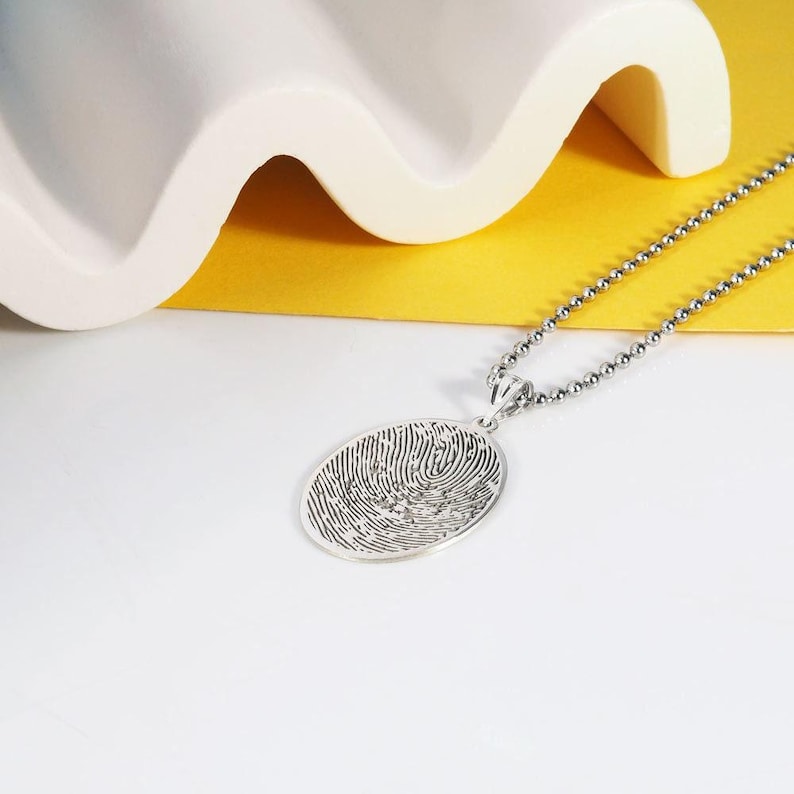 Puede incluir: Un collar de plata con un colgante redondo que presenta un dise&ntilde;o detallado de huella digital. El collar est&aacute; sobre una superficie amarilla y blanca.