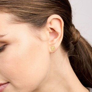May include: Gold-toned number 12 earrings. The earrings are small and delicate, and they are shown being worn in a person's ear.