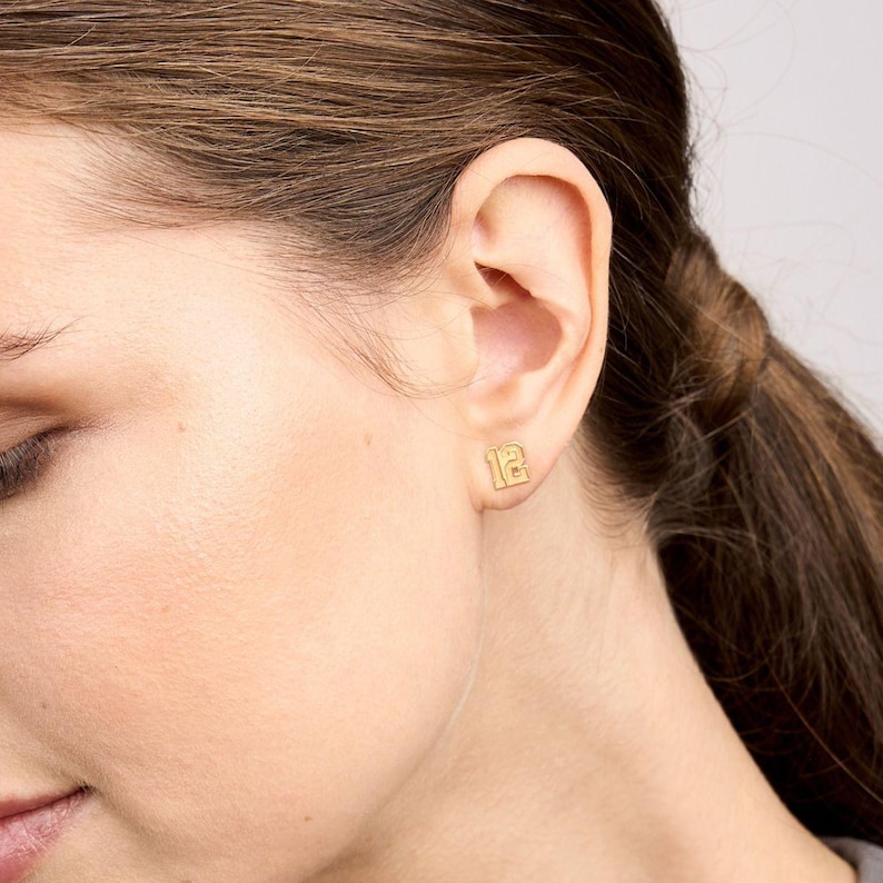 May include: Gold-toned number 12 earrings. The earrings are small and delicate, and they are shown being worn in a person's ear.