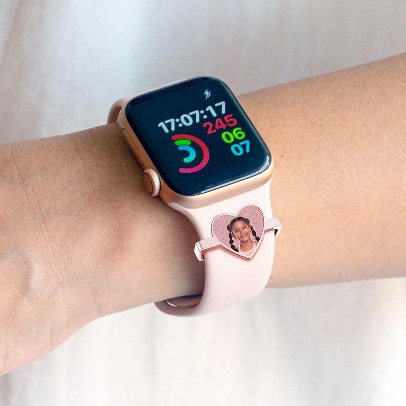 Pu&ograve; includere: Un Apple Watch rosa con un portafoto a forma di cuore che presenta una bambina. Il quadrante dell'orologio mostra l'ora 17:07:17 e un anello di attivit&agrave; colorato.