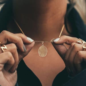 Puede incluir: Un collar de cadena de oro con un colgante ovalado que presenta un dise&ntilde;o detallado de huella digital.