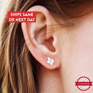 May include: Silver stud earrings featuring a small outline of the state of Texas with the words "Texas Tech" inscribed inside. The earrings are worn in a person's ear.