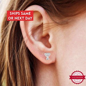 May include: A silver stud earring in the shape of the letter "T" is shown on an ear. The earring is a licensed product. The text "SHIPS SAME OR NEXT DAY" is in a red box in the upper left corner.