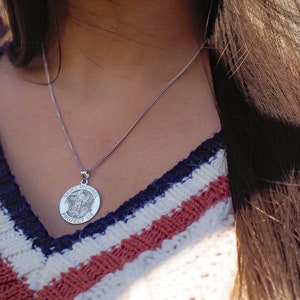 May include: A silver necklace with a round pendant featuring the words "Saint Michael Protect Us". The pendant has a detailed image of an angel with wings.