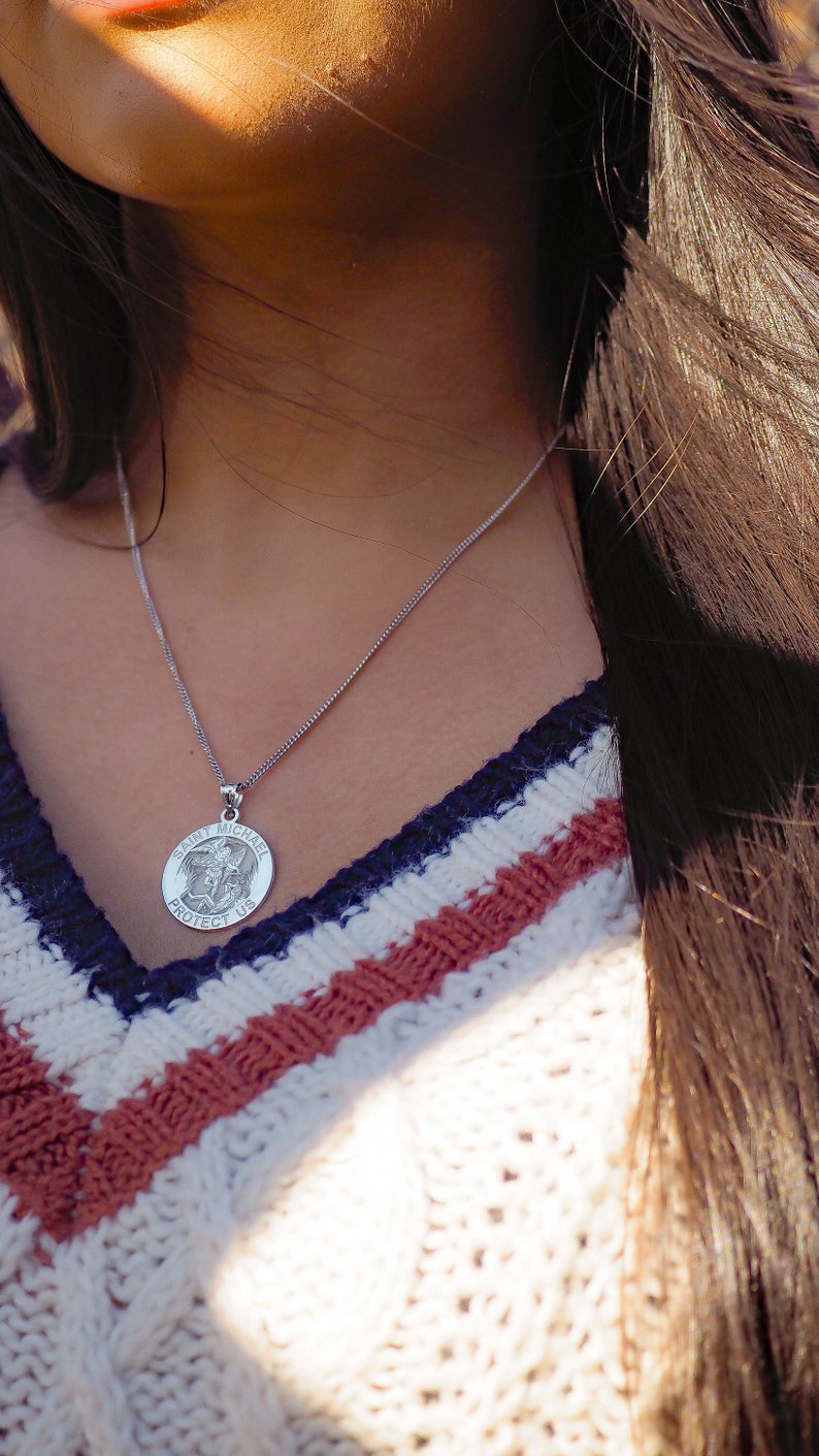 May include: A silver necklace with a round pendant featuring the words "Saint Michael Protect Us". The pendant has a detailed image of an angel with wings.