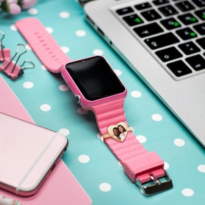 Pu&ograve; includere: Uno smartwatch rosa con uno schermo nero e un ciondolo a forma di cuore con una foto di una coppia. L'orologio &egrave; su uno sfondo blu e bianco a pois.