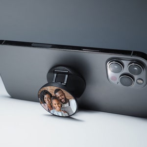 K&ouml;nnte beinhalten: Ein schwarzes Handy-Case mit einem PopSocket, das ein Foto einer dreik&ouml;pfigen Familie zeigt. Der PopSocket ist rund und hat ein schwarz-wei&szlig;es Design. Das Handy-Case ist grau und hat eine Kamera auf der R&uuml;ckseite.