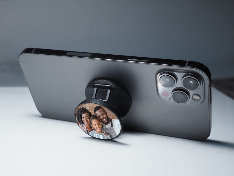 K&ouml;nnte beinhalten: Ein schwarzes Handy-Case mit einem PopSocket, das ein Foto einer dreik&ouml;pfigen Familie zeigt. Der PopSocket ist rund und hat ein schwarz-wei&szlig;es Design. Das Handy-Case ist grau und hat eine Kamera auf der R&uuml;ckseite.