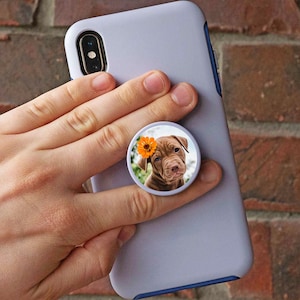 K&ouml;nnte beinhalten: Eine wei&szlig;e Handyh&uuml;lle mit einem Popsocket, das ein braunes Welpen mit einer gelben Blume auf dem Kopf zeigt.