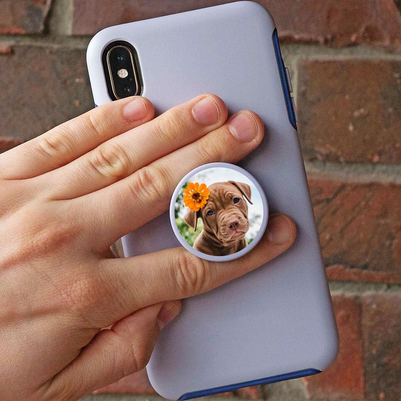 K&ouml;nnte beinhalten: Eine wei&szlig;e Handyh&uuml;lle mit einem Popsocket, das ein braunes Welpen mit einer gelben Blume auf dem Kopf zeigt.