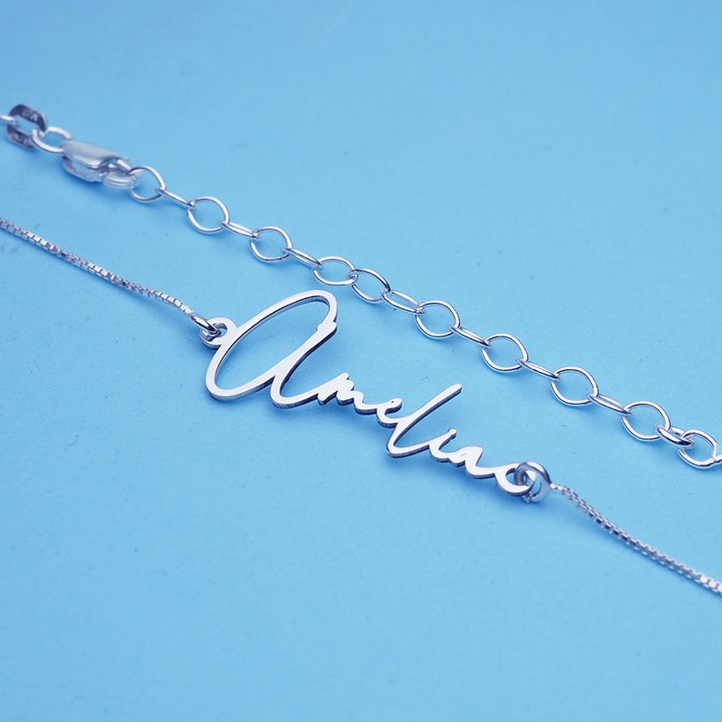 Peut inclure: Un collier en argent avec une cha&icirc;ne d&eacute;licate et un pendentif en forme de plaque cursive qui lit "Amelia".