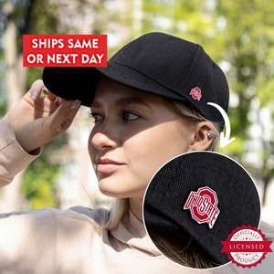 May include: A black baseball cap with a red and white Ohio State pin. The pin features the Ohio State logo and the words "OHIO STATE". The cap is worn by a person.