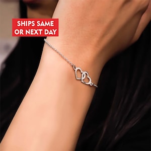 May include: A silver bracelet featuring two intertwined heart pendants. The delicate chain is attached to the hearts, creating a simple yet elegant design. The bracelet is shown on a wrist, with the text "SHIPS SAME OR NEXT DAY" in a red box.
