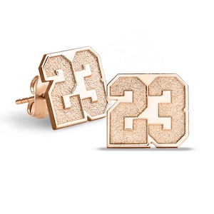 May include: A pair of gold-tone earrings featuring the number 23 in a bold, textured font. The earrings are designed in a square shape with rounded corners.