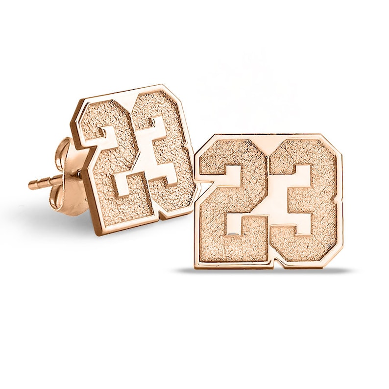 May include: A pair of gold-tone earrings featuring the number 23 in a bold, textured font. The earrings are designed in a square shape with rounded corners.