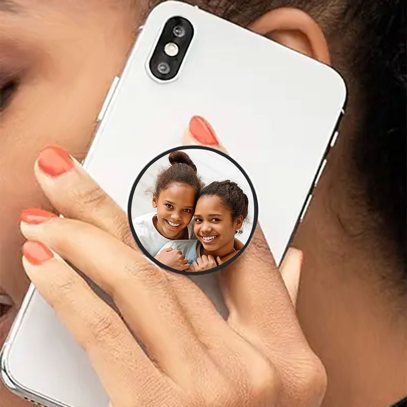 K&ouml;nnte beinhalten: Eine wei&szlig;e Handyh&uuml;lle mit einem schwarzen Handy-Popsocket, auf dem ein Foto von zwei jungen M&auml;dchen zu sehen ist, die in die Kamera l&auml;cheln.