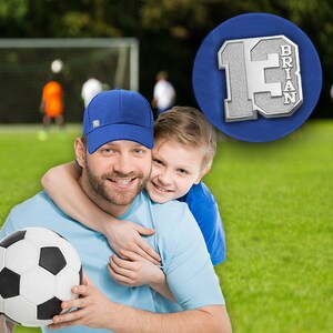 K&ouml;nnte beinhalten: Ein Vater und sein Sohn l&auml;cheln und umarmen sich. Der Vater tr&auml;gt eine blaue Baseballkappe und ein hellblaues Hemd. Der Sohn tr&auml;gt ein blaues Hemd. Sie halten einen Fu&szlig;ball. Das Bild ist vor einem gr&uuml;nen Grasfeld aufgenommen.