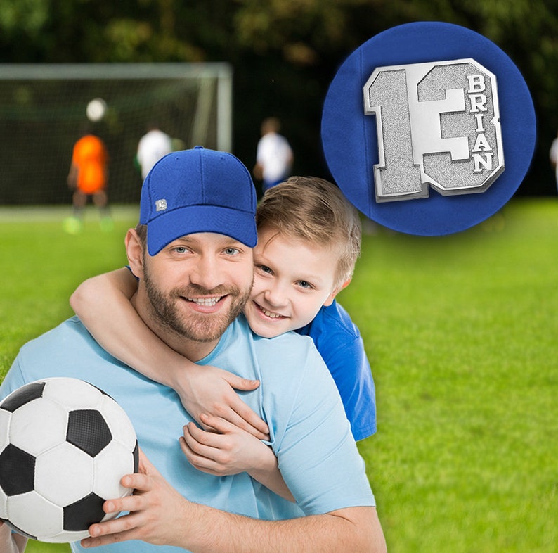 K&ouml;nnte beinhalten: Ein Vater und sein Sohn l&auml;cheln und umarmen sich. Der Vater tr&auml;gt eine blaue Baseballkappe und ein hellblaues Hemd. Der Sohn tr&auml;gt ein blaues Hemd. Sie halten einen Fu&szlig;ball. Das Bild ist vor einem gr&uuml;nen Grasfeld aufgenommen.