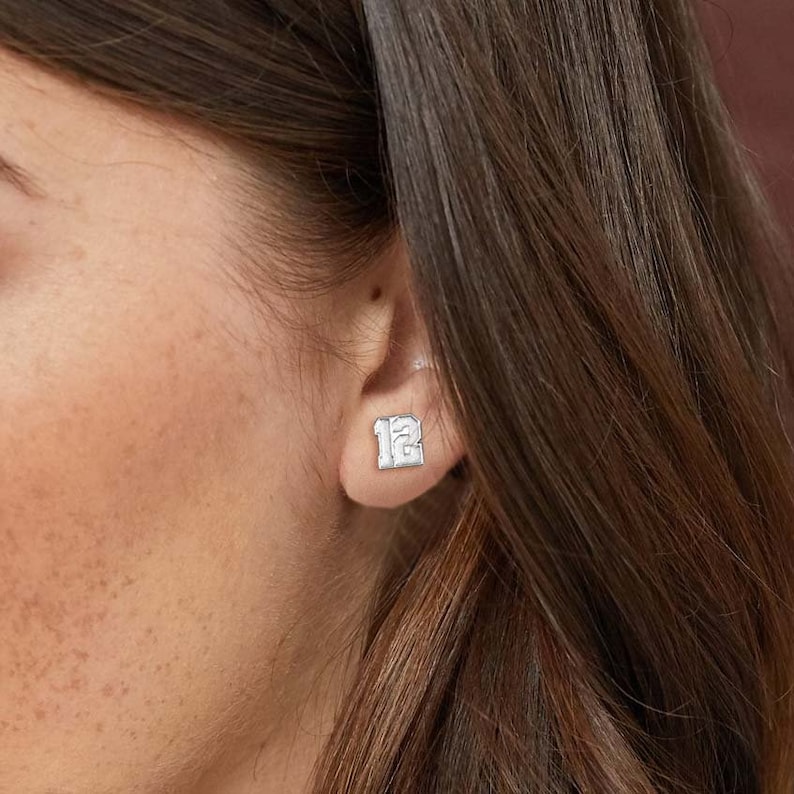 May include: Silver stud earrings with the number "12" engraved on them. The earrings are worn in a person's ear.