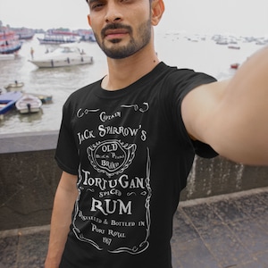 May include: A black t-shirt with a white graphic design of a rum bottle label. The label reads "Captain Jack Sparrow's Old Black Pearl Brand Tortugan Spiced Rum Distilled & Bottled in Port Royal 1967".