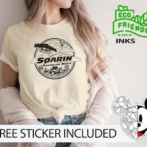 May include: A cream-colored t-shirt with a black and white graphic of a ride called "Soarin' Around the World" at Walt Disney World. The text "Eco Friendly Inks" is in green on a white background.