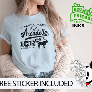 May include: Light blue t-shirt with a black graphic design that says "Finest Quality Arendelle Ice Co. Since 2013". The graphic also includes a stylized image of a moose and crossed pickaxes. The t-shirt is being modeled by a person. The text "ECO FRIENDLY INKS" is in a green box with a leaf design.