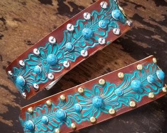 May include: Two brown leather belts with turquoise and silver floral designs and gold and silver studs. The belts are on a wooden surface.  NEZ BLASS