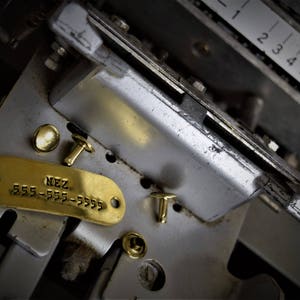 May include: A close-up of a metal machine with a gold-colored metal tag that reads "NEZ 555-555-5555". There are three small gold-colored metal rivets on the machine.