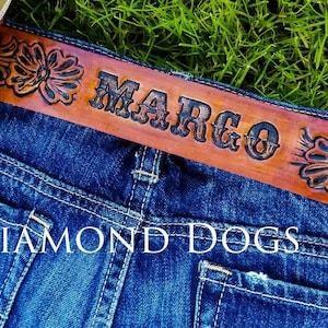 May include: A brown leather belt with a floral design and the name "MARGO" embossed in black lettering. The belt is worn on a pair of blue jeans with the text "THE DIAMOND DOGS" visible.