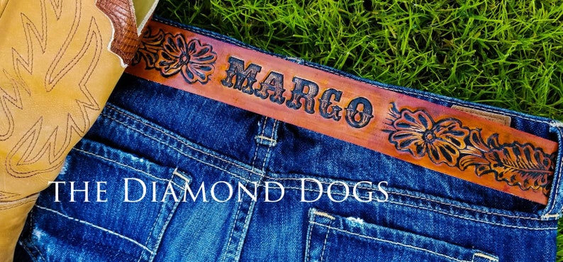 May include: A brown leather belt with a floral design and the name "MARGO" embossed in black lettering. The belt is worn on a pair of blue jeans with the text "THE DIAMOND DOGS" visible.