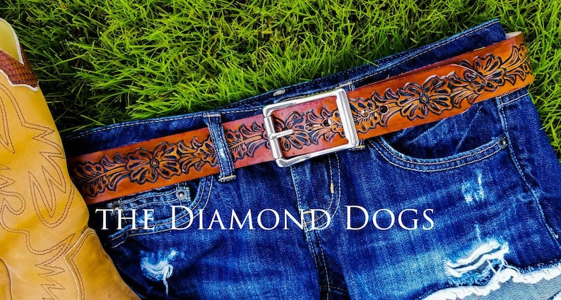 May include: A brown leather belt with a floral design and a silver buckle. The belt is worn on a pair of blue denim shorts. The text "THE DIAMOND DOGS" is visible below the belt.