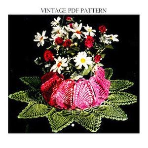 May include: A crocheted doily with a pink crocheted flower pot in the centre. The flower pot is filled with white and pink flowers.