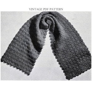 Op de afbeelding: Een grijze gehaakte sjaal met een gegolfde rand. De sjaal is dubbelgevouwen en gedrapeerd over een wit oppervlak.