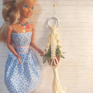 Op de afbeelding: Een pop in een blauwe jurk met sterrenpatroon en een roze riem. Een crèmekleurige macramé plantenhouder houdt een kleine pot met groen vast. De pop heeft blond haar en draagt een ketting.