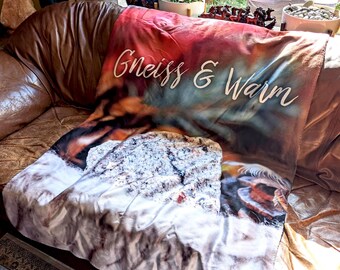 May include: A soft, white and brown fleece blanket with a colorful abstract design and the text "Gneiss & Warm" printed on it. The blanket is folded and draped over a brown leather couch.