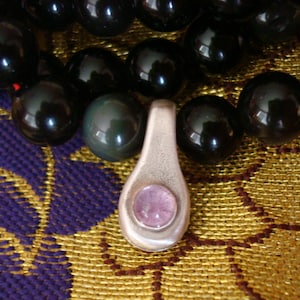 Può includere: Un ciondolo d'argento con una pietra preziosa di colore viola chiaro incastonata al centro, appeso a una collana di perle nere.