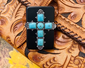 May include: A black rectangular box with a silver cross design featuring turquoise stones. The box is sitting on a brown leather surface with an embossed pattern.