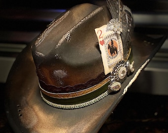 May include: A distressed brown cowboy hat featuring a layered hatband with green and dark red ribbons, a playing card with the text 2 of hearts and Wild Flowers Wild Horses, a silver-toned decorative pin, and a dark feather accent.