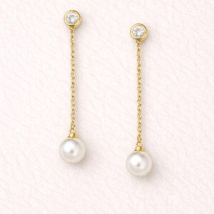 May include: A pair of gold drop earrings featuring a round, bezel-set clear stone at the top, connected by a delicate gold chain to a single white pearl at the bottom. The earrings are designed with a post back and a dangling, linear silhouette.