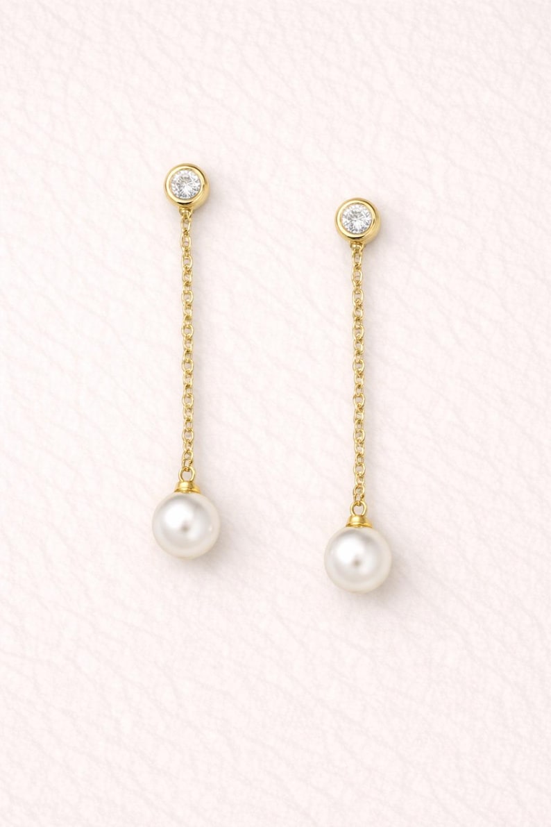 May include: A pair of gold drop earrings featuring a round, bezel-set clear stone at the top, connected by a delicate gold chain to a single white pearl at the bottom. The earrings are designed with a post back and a dangling, linear silhouette.